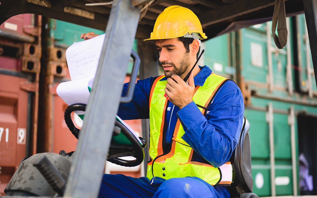 A man on a forklift checks paperwork at a dockyard.